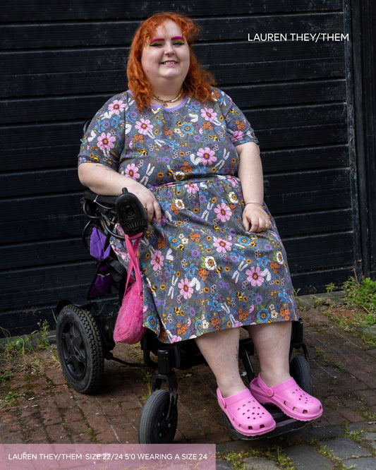 Lauren, a non binary model, with long curly ginger hair and brightly coloured makeup wearing the cottagecore smock dress with pink crocs. They are sat in their mobility aid with pink detailing. The dress print features dragonflies, bees, florals and butterflies on a denim blue base.