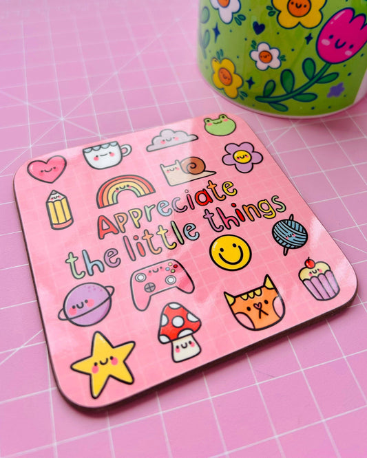 The Appreciate The Little Things drinks coaster by Kelly Lou, sold by Rebel Romance. This square pink coaster features cheerful doodles such as a frog, rainbow, flowers, mushrooms and a smiley face, with the positive quote in colourful lettering. A bright floral mug sits nearby on a pink grid surface.