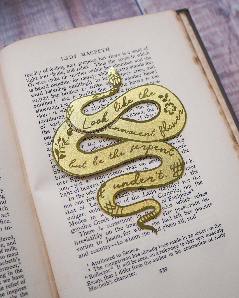 A brass bookmark shaped like a coiled serpent, delicately etched with the famous Lady Macbeth quote: “Look like the innocent flower, But be the serpent under ‘t.” The bookmark is placed inside an open book on a wooden surface, with the title “Lady Macbeth” visible on the page. The snake’s head is designed to peek out from the top of the book when closed, adding a dramatic and literary flair.
