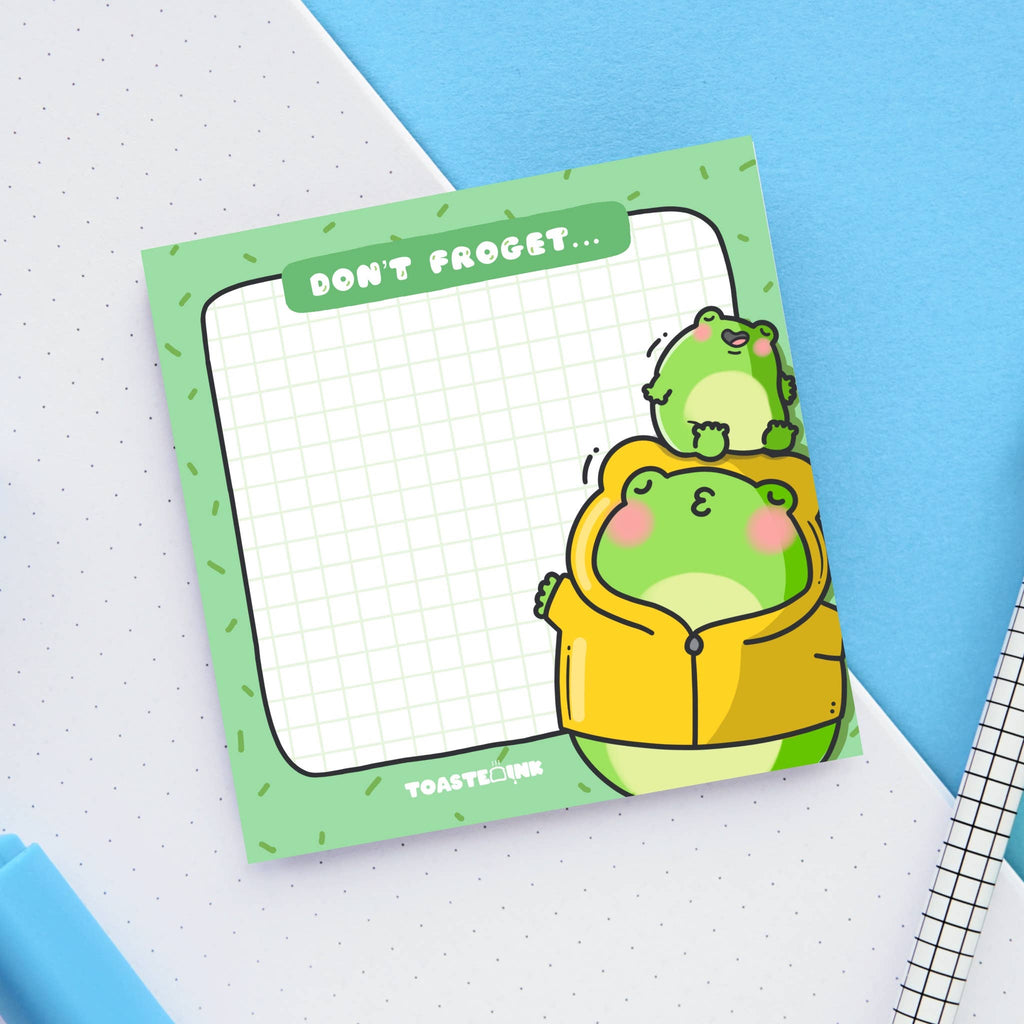 A square frog-themed sticky note pad with a green grid background, featuring a chubby frog in a yellow raincoat being kissed by a smaller frog. The top reads “Don’t Froget…” in a white font on a green tab. Perfect for cute reminders and dopamine-fuelled planning.