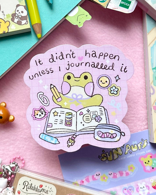 A kawaii journalling sticker designed by Sun Puff Studios, sold by Rebel Romance, a small UK business. The glossy vinyl sticker features a cute pastel frog holding a pencil with the quote “It didn’t happen unless I journalled it.” Surrounded by cheerful icons like a cat, smiley face, and open planner, this pink grid-style sticker is perfect for scrapbooking, planners, and journalling lovers. Flatlay on a pink background with cute stationery.