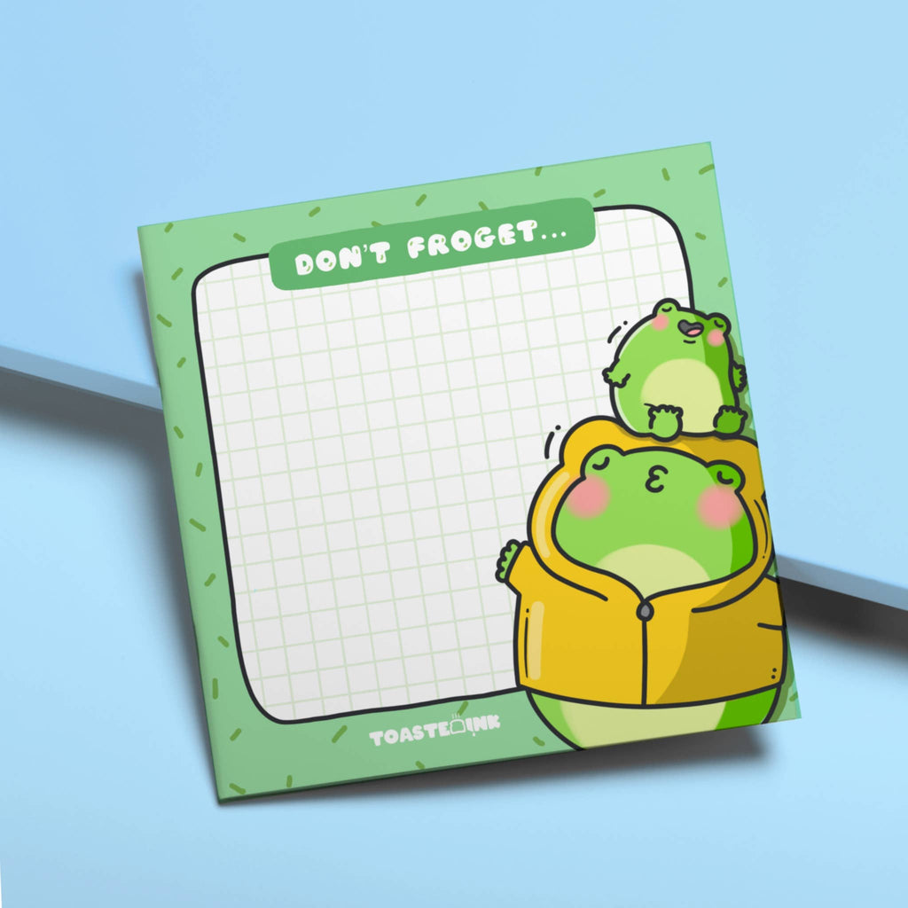 A square frog-themed sticky note pad with a green grid background, featuring a chubby frog in a yellow raincoat being kissed by a smaller frog. The top reads “Don’t Froget…” in a white font on a green tab. Perfect for cute reminders and dopamine-fuelled planning.