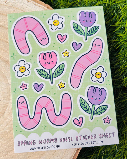 Flat lay of Rebel Romance Spring Worms Vinyl Sticker Sheet by Kelly Lou, featuring cute pink worms, smiling flowers, pastel tulips, and hearts on a green grid background. Printed on waterproof A6 vinyl with a glossy finish, ideal for decorating notebooks, laptops, or crafting. A whimsical spring-themed stationery treat for millennial lovers of quirky, kawaii designs.