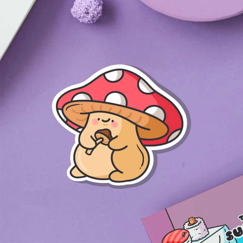 A cute cartoon-style vinyl sticker of a chubby tan mushroom character with a red and white spotted cap, holding a smaller mushroom in its hands. Displayed on a blue background beside a teacup and flower. Waterproof, glossy finish – ideal for decorating everyday items.