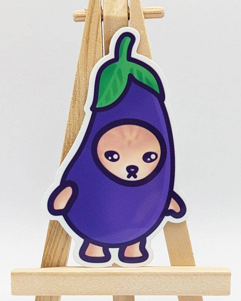 Rebel Romance Eggplant Cat meme vinyl sticker by Mona Mauve, shown upright on a wooden mini easel. The waterproof sticker features a cartoon cat in a purple aubergine costume with a leafy green top. From the Snack Cats collection, this cute and slightly melancholic design is perfect for gifting, journaling, or adding a touch of food-themed fun to your accessories. Ideal for kawaii lovers and alternative style fans.