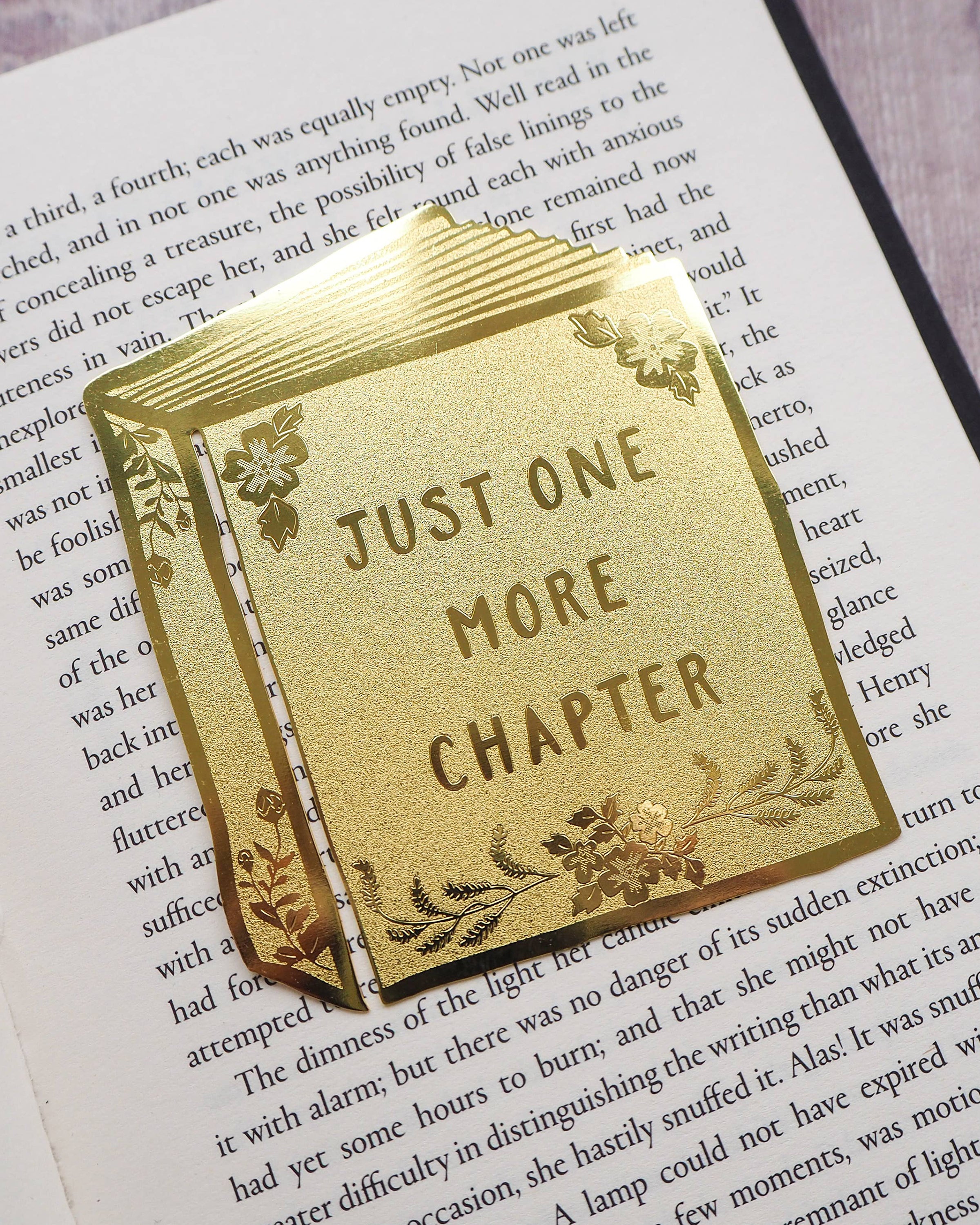 A shiny gold brass bookmark shaped like an open book with floral details and the words “Just One More Chapter” on it, by Literary Emporium. Styled on an open novel, sold by Rebel Romance, a UK small business celebrating alternative style, gifts and slow ethical fashion.