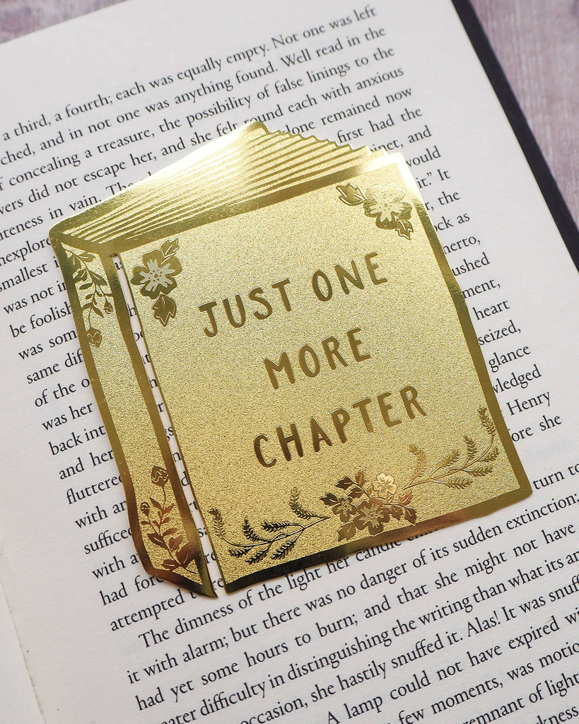 A shiny gold brass bookmark shaped like an open book with floral details and the words “Just One More Chapter” on it, by Literary Emporium. Styled on an open novel, sold by Rebel Romance, a UK small business celebrating alternative style, gifts and slow ethical fashion.