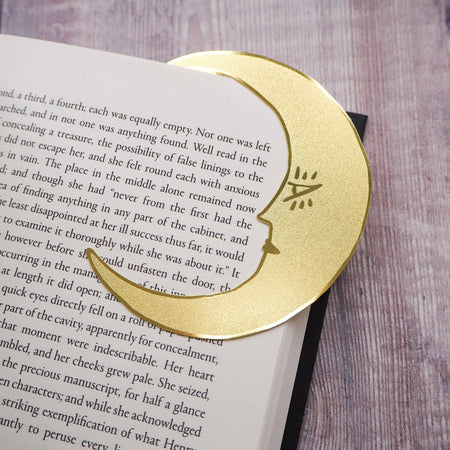 A crescent moon-shaped brass bookmark with “Midnight Readers Club” and celestial stars etched onto its surface, placed on an open book to mark a page. The bookmark adds an elegant and whimsical touch, ideal for book lovers who enjoy night-time reading.