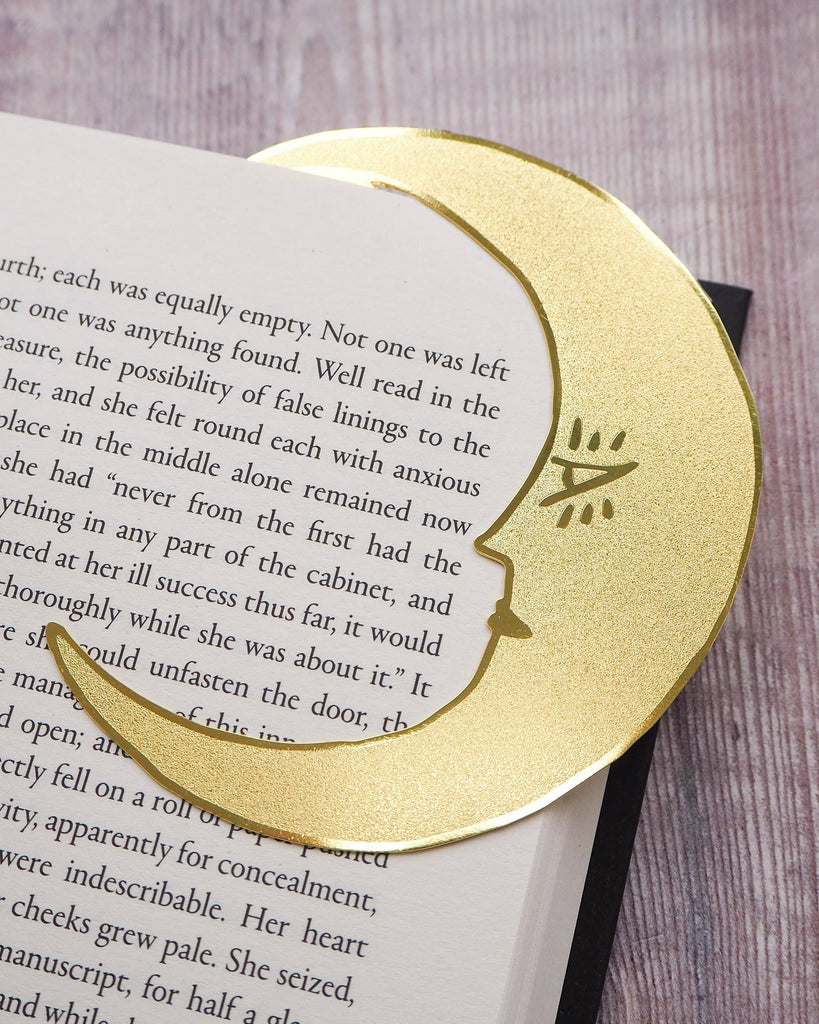 A crescent moon-shaped brass bookmark with “Midnight Readers Club” and celestial stars etched onto its surface, placed on an open book to mark a page. The bookmark adds an elegant and whimsical touch, ideal for book lovers who enjoy night-time reading.