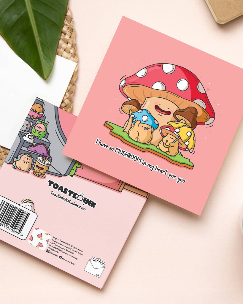 Flat lay photo of a square pink greetings card by Toastedink, stocked at Rebel Romance. The card features cute cartoon mushrooms with smiling faces, hugging each other under a red and white spotted toadstool. Text reads “I have so MUSHROOM in my heart for you.” A whimsical and punny design for gifting mushroom lovers or sending heartfelt notes.