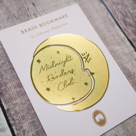 A crescent moon-shaped brass bookmark with “Midnight Readers Club” and celestial stars etched onto its surface, placed on an open book to mark a page. The bookmark adds an elegant and whimsical touch, ideal for book lovers who enjoy night-time reading.