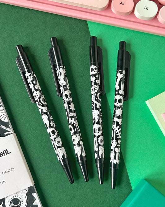 Flat lay of four black ballpoint pens with white skulls, bones, ghosts and stars, next to a gothic notebook and pastel keyboard. These spooky pens are by Rebel Romance, a UK indie brand selling alternative stationery and gifts.