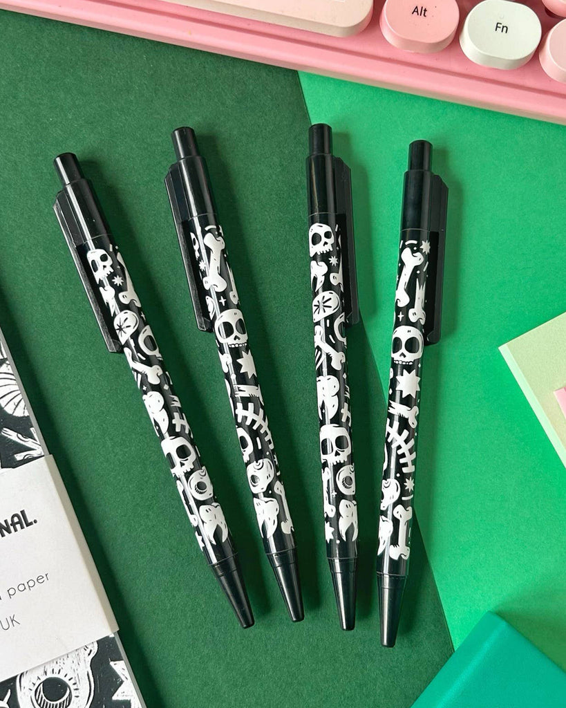 Flat lay of four black ballpoint pens with white skulls, bones, ghosts and stars, next to a gothic notebook and pastel keyboard. These spooky pens are by Rebel Romance, a UK indie brand selling alternative stationery and gifts.