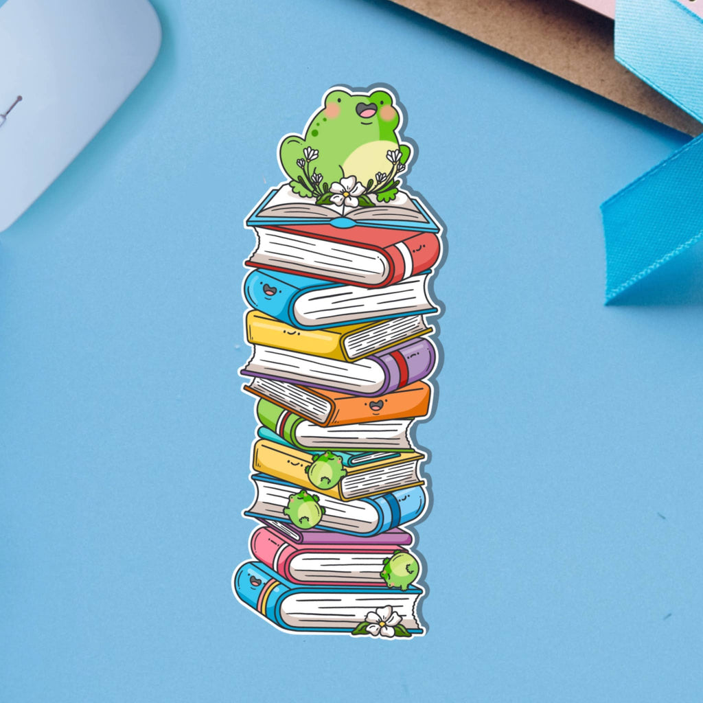 A cute rectangular bookmark showing a cheerful green frog sitting on top of a tall, colourful stack of cartoon books with smiling faces. Printed on glossy card with flowers and tiny frogs tucked between the books. A playful accessory for readers and stationery lovers.