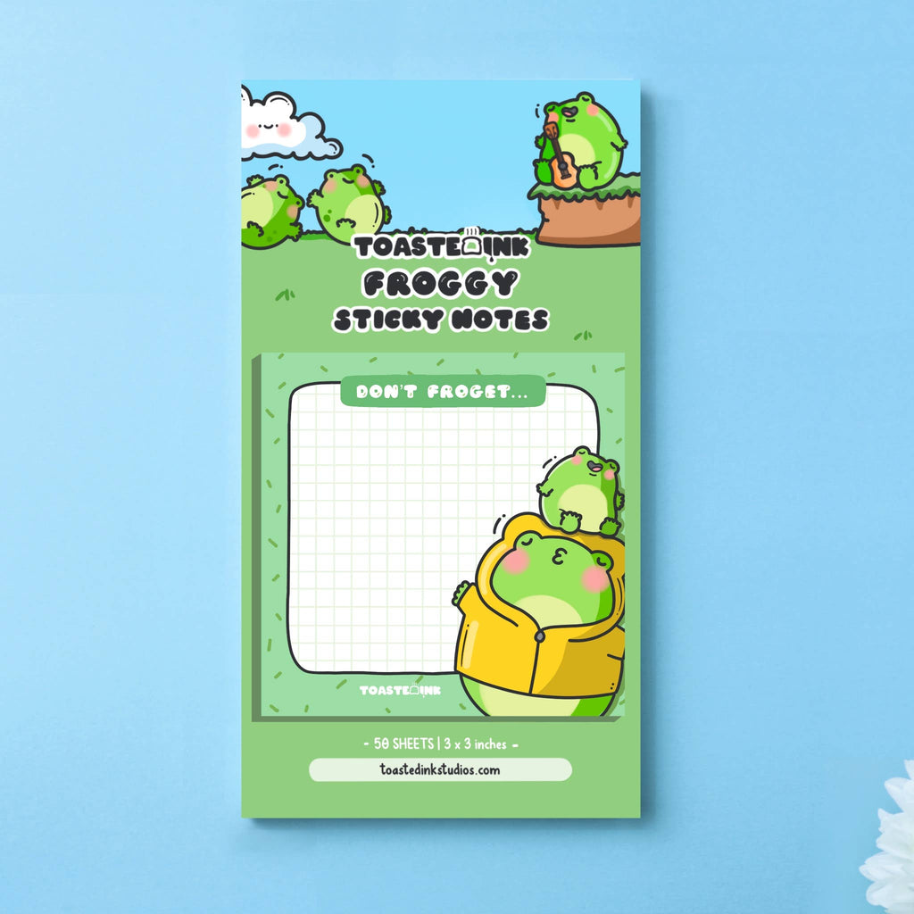 A square frog-themed sticky note pad with a green grid background, featuring a chubby frog in a yellow raincoat being kissed by a smaller frog. The top reads “Don’t Froget…” in a white font on a green tab. Perfect for cute reminders and dopamine-fuelled planning.