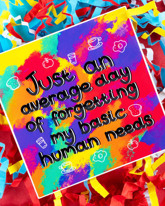Rainbow-splattered vinyl sticker with the phrase “Just an average day of forgetting my basic human needs,” featuring cute white food doodles. The sticker is laid on top of bright red, blue, and yellow shredded confetti. Sold by Rebel Romance, a UK-based small business known for alternative, inclusive fashion and quirky gifts for millennials.