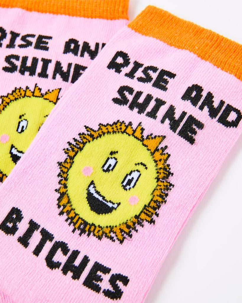 Close-up of two pink socks with an orange cuff and bold black lettering that says "Rise and Shine Bitches" with a happy sunshine face in the middle. Stocked by Rebel Romance, a UK small business for alternative gifts and retro-themed goodies.
