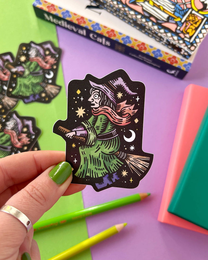 A hand with green painted nails holds a vinyl sticker of a witch flying on a broomstick against a black starry background. The witch has a green dress, purple hat, and red scarf, with a crescent moon and stars around her. In the background are more witch stickers, green and yellow pencils, and colourful books. A vibrant flat lay by Rebel Romance, a UK brand known for alternative gifts and slow fashion.