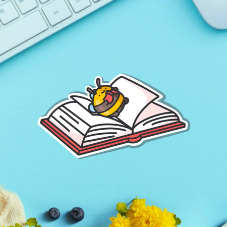A cute vinyl sticker of a yellow and brown bumblebee lying on its back on top of an open book, tongue sticking out playfully. Displayed on a purple background with pink and blue elements. Glossy waterproof finish, ideal for book lovers and readers.
