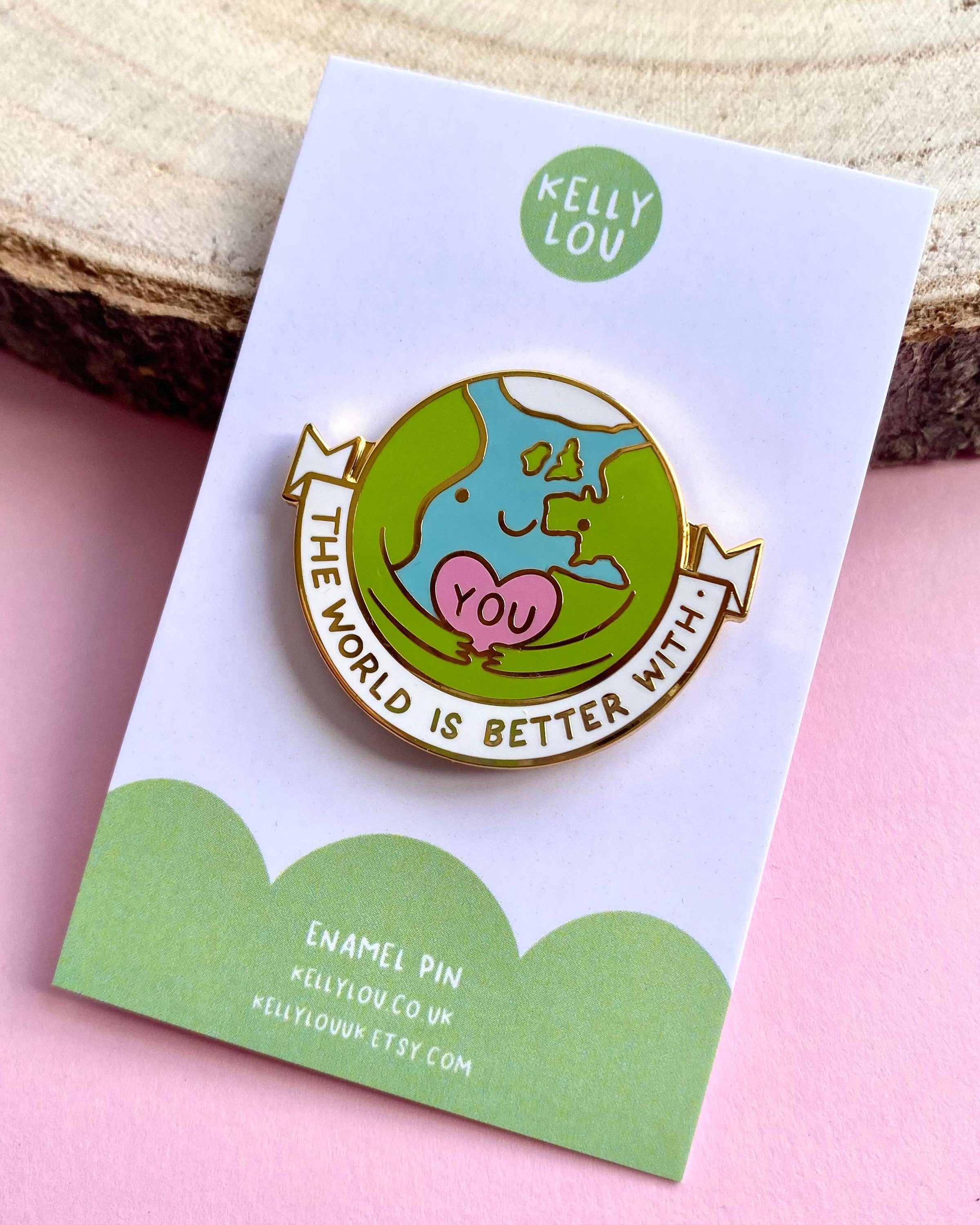 A close-up of The World Is Better With You hard enamel pin by Kelly Lou, displayed on its backing card against a soft pink background and wooden slice. Sold by Rebel Romance, a UK brand for millennials who love alternative style and positive mental health designs. The pin features a cute Earth cuddling a pink heart that says “YOU.”