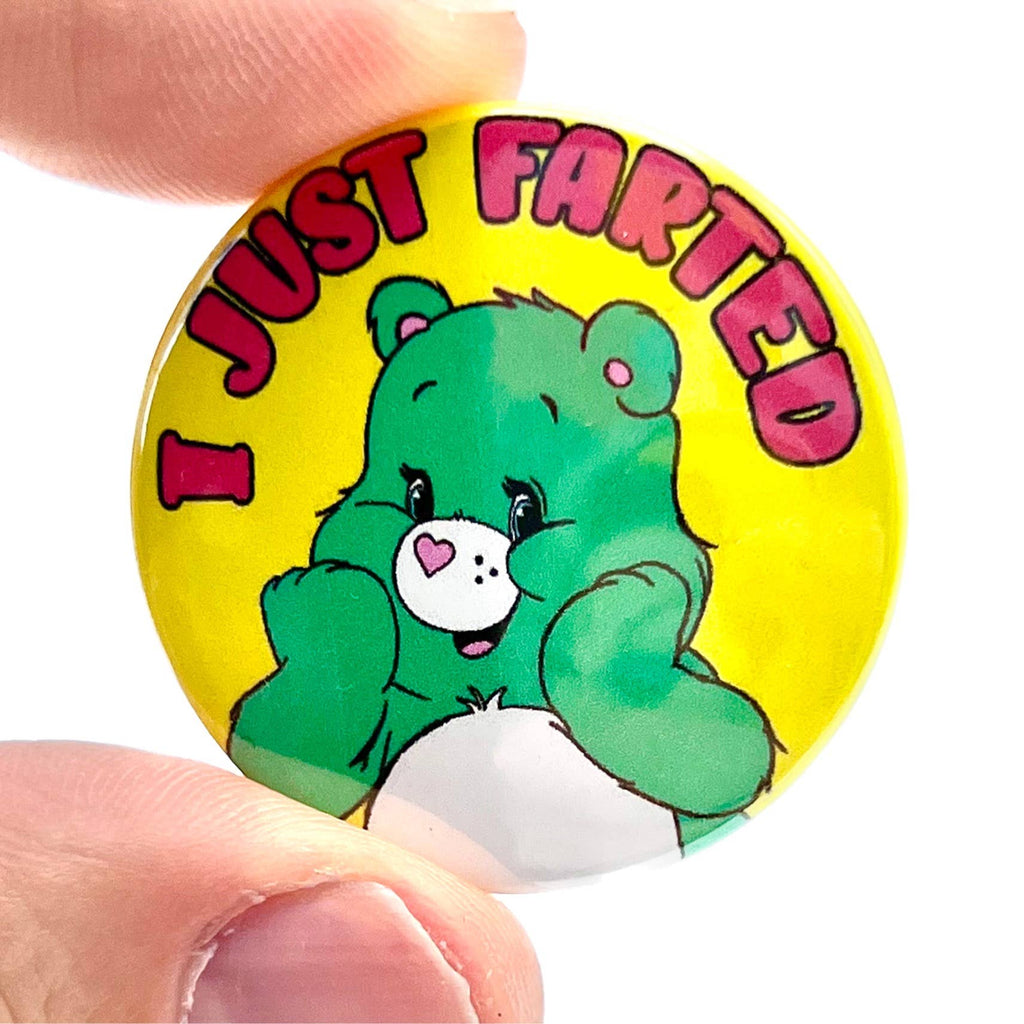 A person holding a small round pin badge with a vintage-style green cartoon bear blushing and covering its cheeks. The badge has bold red and pink text reading “I JUST FARTED” on a yellow background. Designed by Bite Your Granny and available from Rebel Romance, a UK indie brand specialising in quirky, fun, and nostalgic fashion accessories and gifts for alternative millennials.