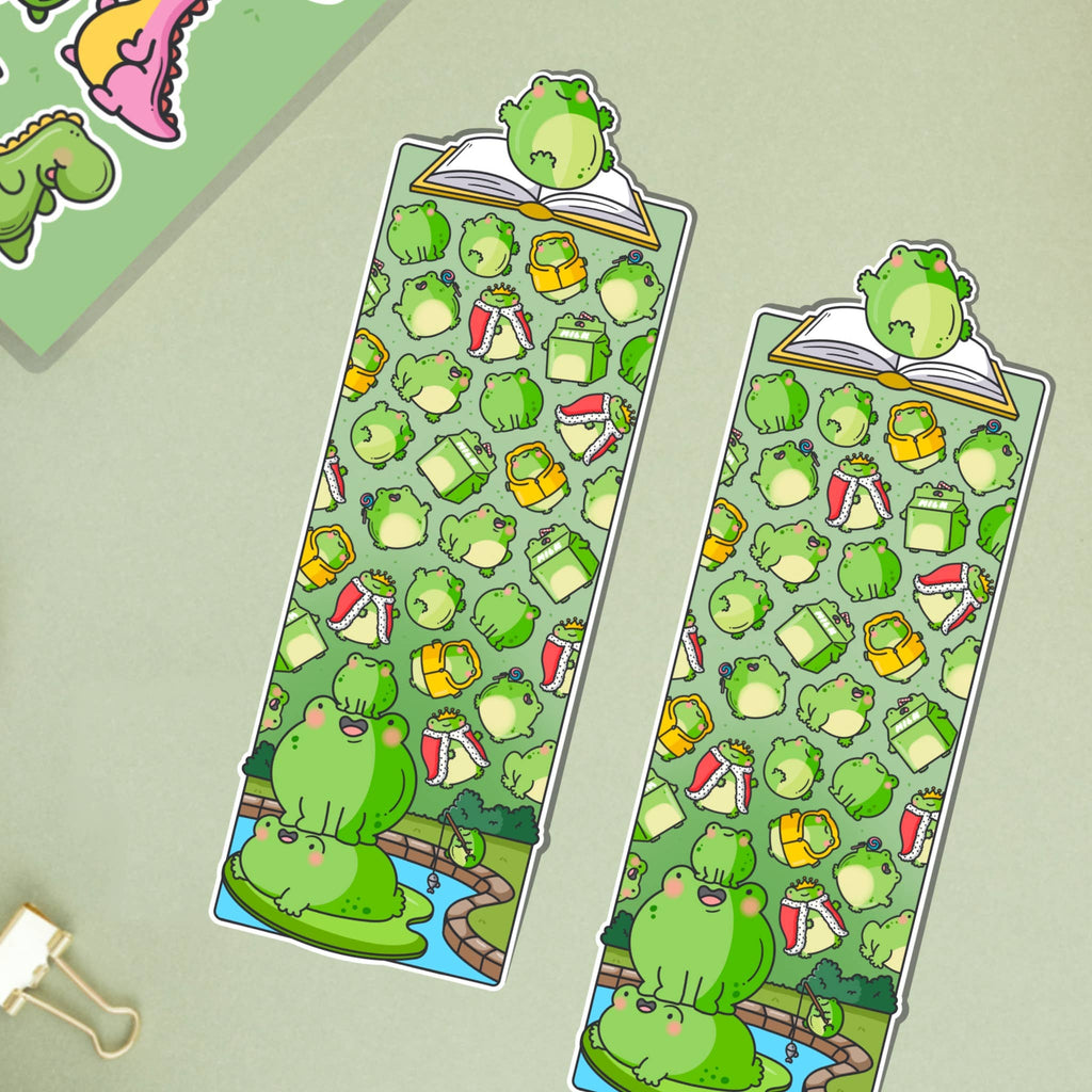 Glossy die-cut bookmark featuring a colourful pattern of cartoon frogs, drinks, books, and accessories on a green background. A frog sits atop an open book at the top, with more frogs stacked on a lily pad at the bottom.