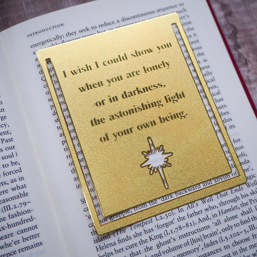 A close-up image of a brass bookmark engraved with the heartfelt Hafiz of Shiraz quote: “I wish I could show you, when you are lonely or in darkness, the astonishing light of your own being.” The bookmark is resting on the pages of an open book, showcasing its intricate detailing and elegant design.