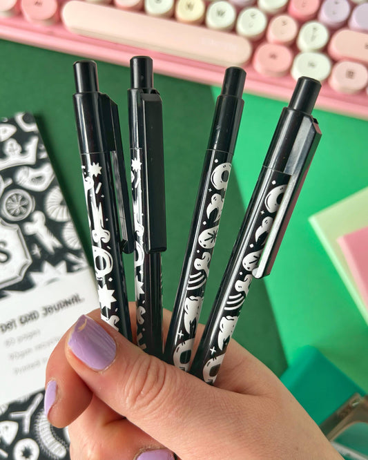 Hand holding four black ballpoint pens with spooky white skull and bones illustrations, perfect for gothic stationery fans. The pens are made by Rebel Romance, a UK small business known for unique, alternative gifts and slow fashion.
