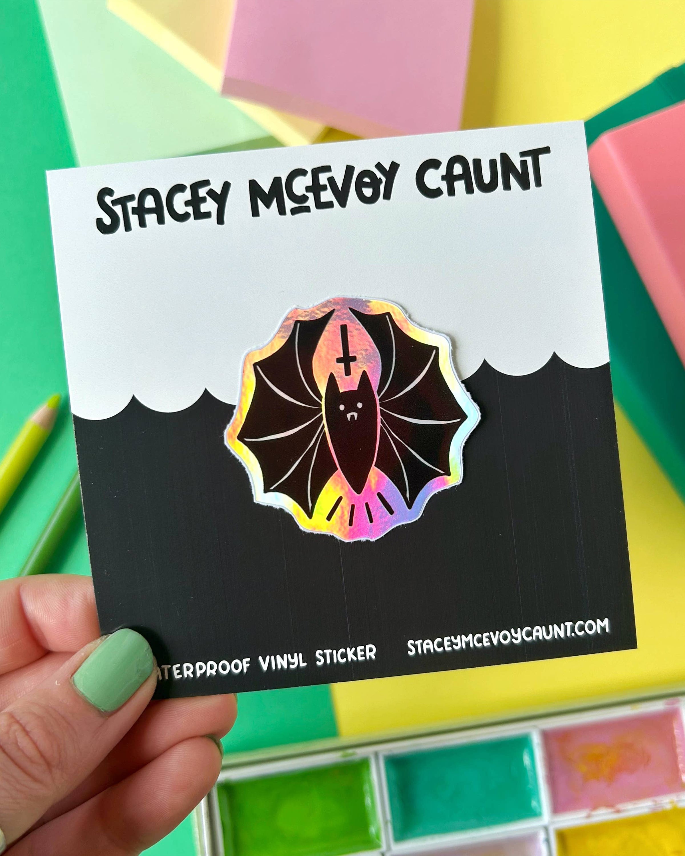 Close-up of a hand holding a rainbow holographic bat sticker mounted on a branded Stacey McEvoy Caunt backing card. The sticker features a black vampire bat with a cute face, fangs, and a gothic upside-down cross above its head. Set against a colourful flat lay with pastel notebooks and art supplies, perfect for showcasing alternative accessories by Rebel Romance.