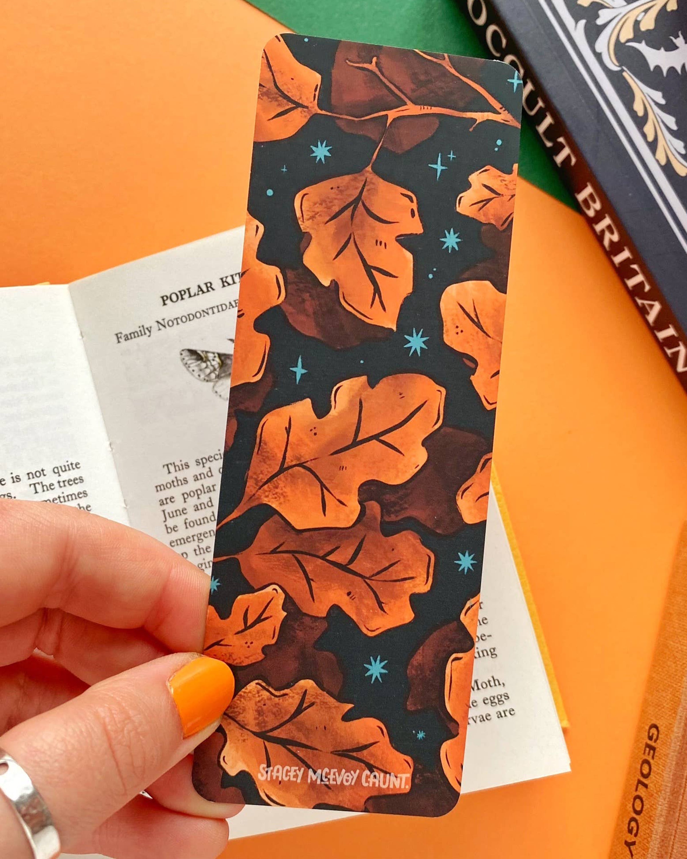 Close-up of a hand holding a Rebel Romance Autumn Leaves Bookmark in front of an open book. The bookmark features orange and brown illustrated leaves on a dark background with small teal stars, and has a matte velvet texture. Books and orange paper are visible in the background.