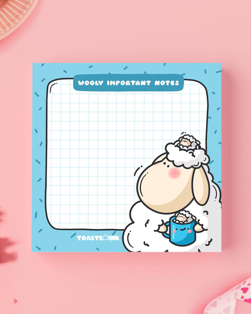 Toastedink sticky note pad from Rebel Romance featuring a cute sheep holding a mug on a fluffy cloud. Blue border with sprinkles, grid paper centre, and the words “Wooly Important Notes”. Displayed on a pastel pink background with stationery.