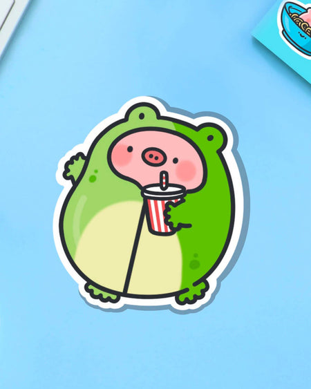 Quirky vinyl sticker of a pig in a frog costume holding a stripy drink, styled on a pastel blue backdrop with office items. A fun addition to your desk or tech. Designed by Toastedink, available at Rebel Romance – a UK brand embracing alternative, creative living.