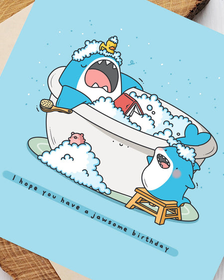 Close-up of Toastedink's shark birthday card design. A shark reads in the bath while another shark tries to wake him. Cute and quirky greeting card sold by Rebel Romance, perfect for birthdays with a splash of fun.