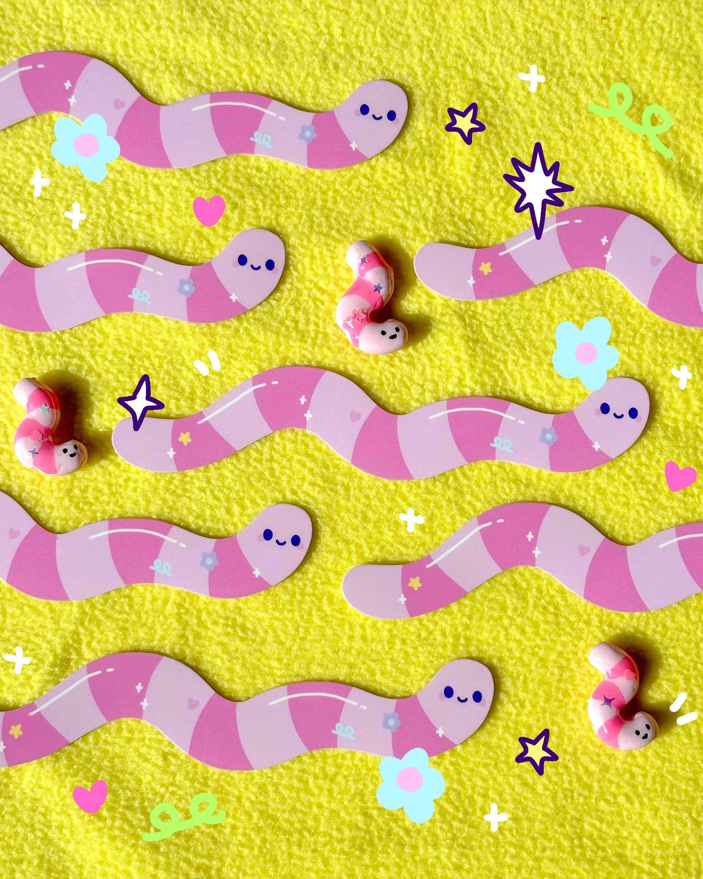 Cute pastel pink striped worm stickers with smiling faces arranged on a bright yellow textured background. The Long Wormie Stickers by Sun Puff Studios are surrounded by clay worm figures and digital doodles of flowers, stars, and hearts. Designed for Rebel Romance, a small UK brand celebrating alternative fashion and millennial aesthetics with handmade and quirky gifts.