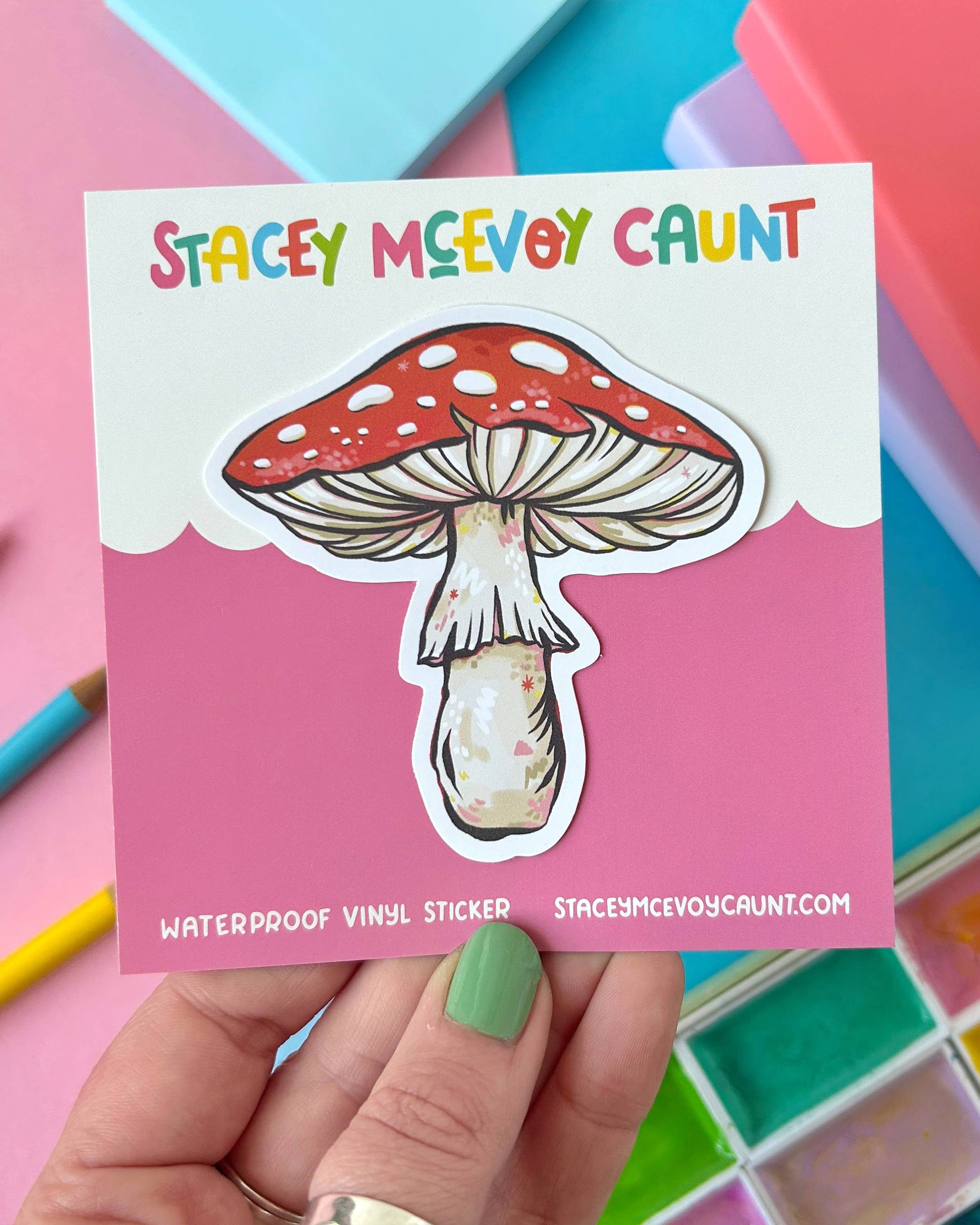 A colourful vinyl sticker of a red and white toadstool mushroom, shown on a pink branded backing card that reads “Stacey McEvoy Caunt.” The sticker is waterproof and professionally printed, held by a hand with green painted nails. Perfect for laptops, notebooks or water bottles. Sold by Rebel Romance, a UK small business celebrating alternative aesthetics and quirky gifts.