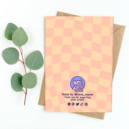 Back of a peach and orange checkered anniversary card designed by Mona Mauve for Rebel Romance. Features a cute cat logo with a QR code and the message “Made by @mona_mauve – Thank you for supporting small artists!” Social media icons for Etsy, Instagram, Twitter, TikTok, and Pinterest are shown at the bottom. Card is displayed with a brown kraft envelope and eucalyptus sprig on a white background.