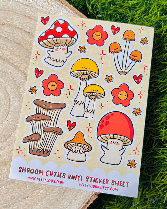 Flat lay photo of the Shroom Cuties Vinyl Sticker Sheet by Kelly Lou, available at Rebel Romance. The sheet features cute cartoon mushrooms with smiling faces in red, yellow, orange, and brown, plus hearts and flowers, on a beige grid background. Fun and waterproof, these stickers are perfect for decorating journals, laptops, or gifts. Shot on a wooden log slice with grass background.