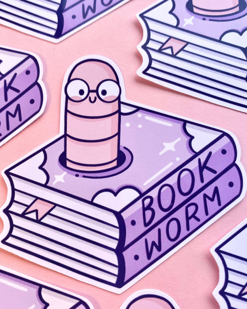 Rebel Romance Bookworm Glossy Sticker by Kelly Lou shown on a pink background, with a smiling worm wearing glasses emerging from a purple book decorated with hearts and sparkles. The word “BOOKWORM” is written on the spine. A kawaii vinyl sticker perfect for laptops, book journals, or gifting to fellow bookish friends.