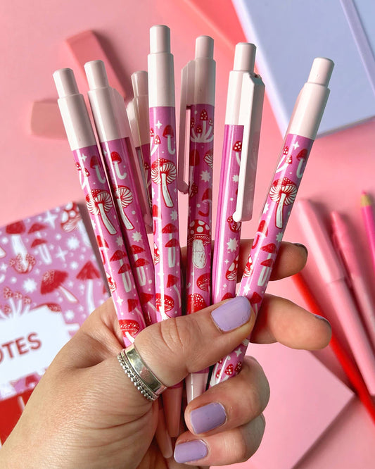 A handful of Rebel Romance’s pink mushroom pens fanned out in a hand with lavender nails. These ballpoint pens feature a magical mushroom print designed by Stacey McEvoy Caunt, ideal for cottagecore and stationery fans.
