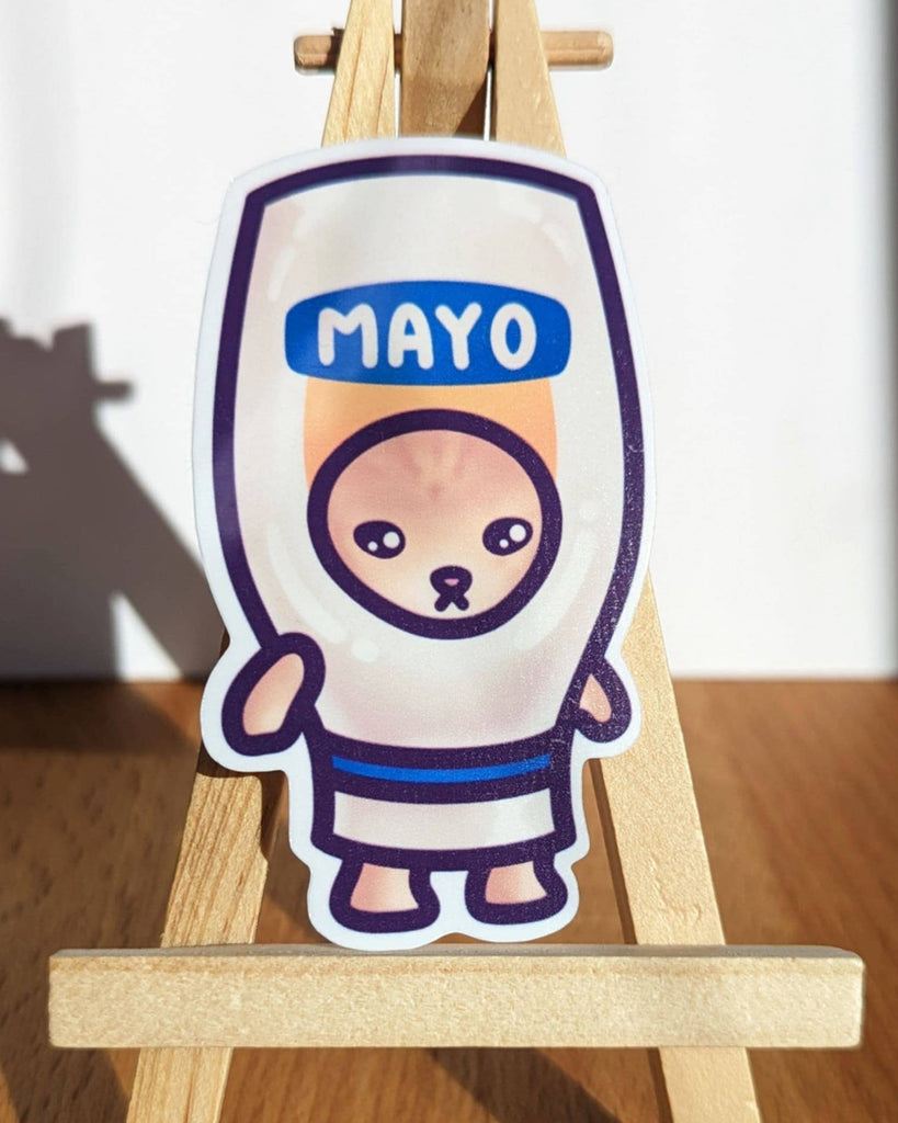 Close-up of Rebel Romance Mayo Cat meme vinyl sticker by Mona Mauve, displayed upright on a mini wooden easel. Features a sad cartoon cat dressed as a squeezable mayo bottle with bold “MAYO” text. From the Snack Cats collection, this waterproof and durable sticker is ideal for alternative style lovers, kawaii collectors, and food-themed stationery fans.