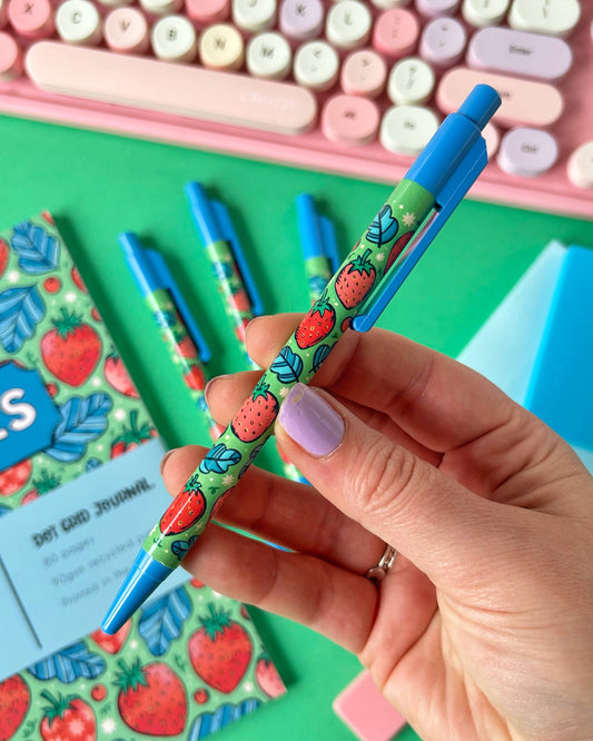 A close-up of a hand holding the Rebel Romance strawberry ballpoint pen against a colourful desk setup. The pen features a green background with strawberries and leaves, a bright blue clicker top and nib, and a matching notebook in the background.