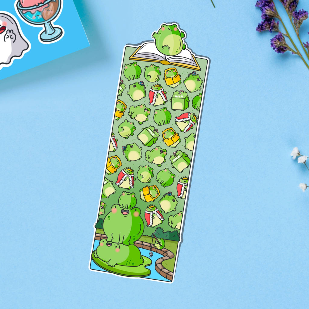Glossy die-cut bookmark featuring a colourful pattern of cartoon frogs, drinks, books, and accessories on a green background. A frog sits atop an open book at the top, with more frogs stacked on a lily pad at the bottom.