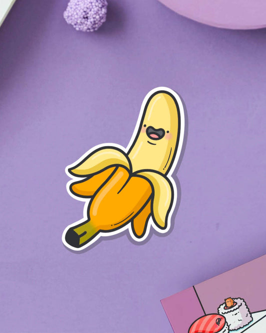 Toastedink banana sticker at Rebel Romance, featuring a smiling cartoon banana with a peeled design. Photographed on a soft purple backdrop with cute accessories nearby.