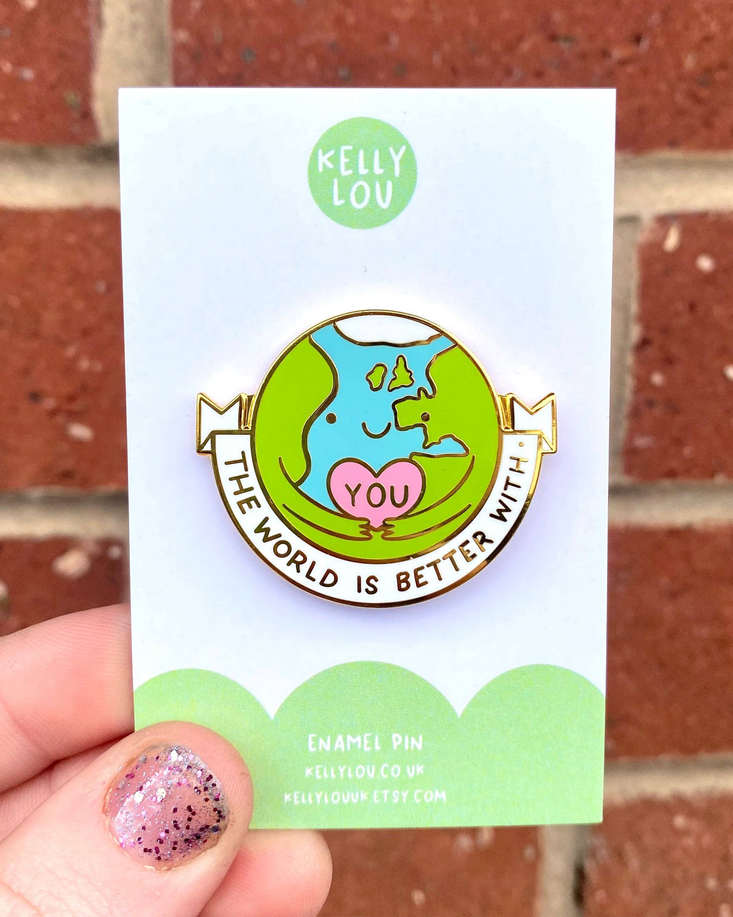 A close-up of The World Is Better With You hard enamel pin by Kelly Lou, photographed against a red brick wall. Sold by Rebel Romance, a UK small business celebrating alternative fashion and gifts for millennials. The pin shows a smiling Earth hugging a pink heart with “YOU,” with the words “The World Is Better With” around it.