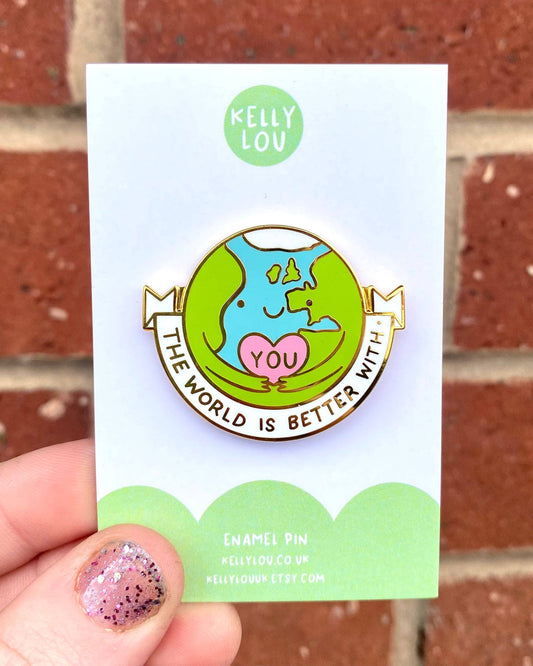 A close-up of The World Is Better With You hard enamel pin by Kelly Lou, photographed against a red brick wall. Sold by Rebel Romance, a UK small business celebrating alternative fashion and gifts for millennials. The pin shows a smiling Earth hugging a pink heart with “YOU,” with the words “The World Is Better With” around it.
