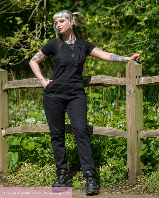 An alternative non binary femme model, Faeryn, wearing the rebel romance black dungarees with  black short sleeve crop top and black platform trainers. They are stood outside leaning on a wooden fence whilst looking left and a green park behind them. 