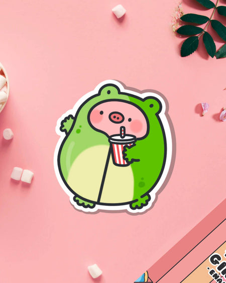 Toastedink vinyl sticker showing a chubby pig dressed in a frog outfit holding a drink, styled on a soft pink background with mini marshmallows. A fun and playful design sold by Rebel Romance, a UK indie store for millennials who love cute, retro and alternative aesthetics.