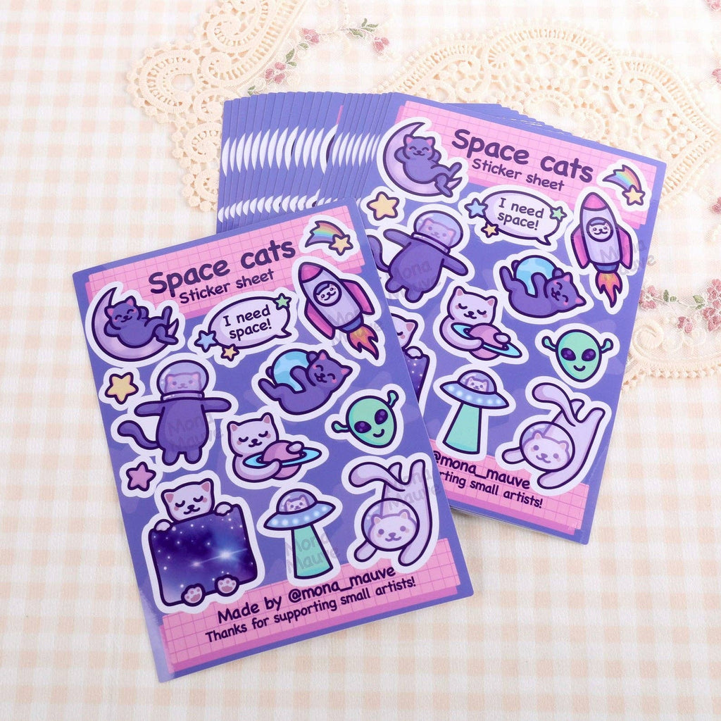 Stack of Space Cats Sticker Sheets by Mona Mauve displayed on a soft, patterned fabric. Each sheet includes 13 vibrant vinyl stickers with space-themed cat illustrations, featuring stars, spaceships, and aliens. A whimsical and fun stationery gift created by a small independent artist.