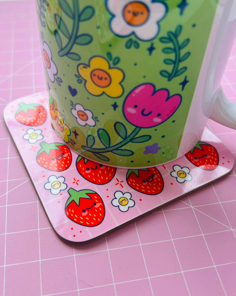 A cute Strawberry Cuties Drinks Coaster by Kelly Lou from Rebel Romance. The pink square coaster features smiling red strawberries and tiny happy white flowers. It’s shown in use under a bright green floral mug, perfect for adding fun, alternative style to your home décor.