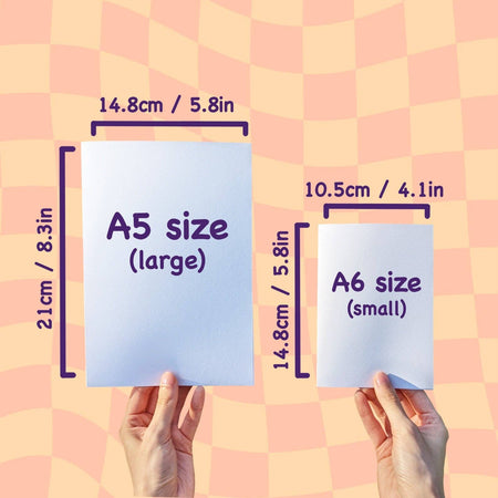 Size comparison chart showing A5 and A6 card dimensions on a peach checkerboard background. One hand holds an A5 card labelled “A5 size (large)” with measurements 14.8cm x 21cm (5.8in x 8.3in), and the other holds an A6 card labelled “A6 size (small)” with measurements 10.5cm x 14.8cm (4.1in x 5.8in). Designed to help customers visualise greeting card sizes from Rebel Romance and Mona Mauve.